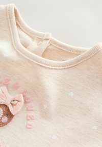 Camisa de algodón beige claro con cuello redondo, diseño bordado de osito con lazo y letras rosas. Tejido texturizado con patrones de corazones.