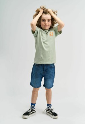 Tricou scurt cu mâneci, de culoare verde deschis, cu un logo portocaliu, purtat cu șorturi din denim bleu, adidași negri și șosete cu carouri albastre. Păr creț.