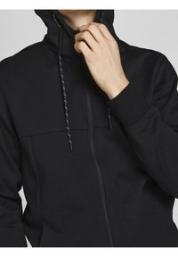 Uomo che indossa una felpa nera con zip e cappuccio, mentre tiene il colletto vicino al mento con la mano destra su uno sfondo semplice.