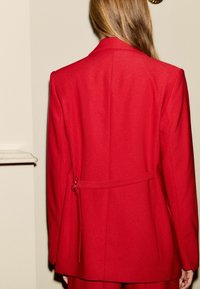 Rød skræddersyet blazer med en semi-fittet form, inkluderer en rygsløjfe og et glat, tekstureret stof, med et klassisk reversdesign.