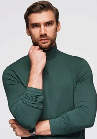 Grüner Rollkragenpullover aus Stretchstoff, mit eng anliegenden Ärmeln und gerippten Details. Getragen mit einer Uhr am Handgelenk.
