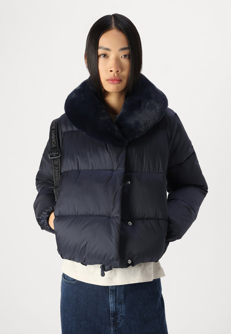 Granatowa kurtka puchowa z kołnierzem z sztucznego futra, zapięciem na napy i elastycznym wykończeniem, z markowym paskiem na jednym ramieniu.