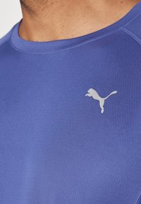 Camisola desportiva azul feita de tecido texturizado, com um logótipo prateado da Puma no lado esquerdo do peito. Decote redondo e mangas curtas.