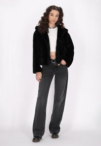 Giacca cropped in finta pelliccia nera con cappuccio, abbinata a jeans neri a vita alta e a un top corto bianco. Accenti in hardware argentato visibili.