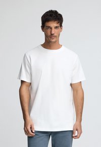 Camiseta de algodón blanca con cuello redondo y mangas cortas, que presenta un diseño minimalista y una pequeña etiqueta de tela en el lado inferior.