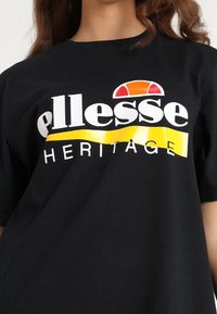 Schwarzes Baumwoll-T-Shirt mit auffälliger weißer Schrift "ellesse" und "HERITAGE" sowie oben einem Logo in Gelb und Rot. Kurzärmelig, lockere Passform.