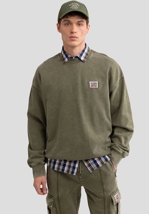 Olijfgroene oversized sweatshirt met een patch, gelaagd over een blauw-wit geruit shirt, in combinatie met bijpassende olijfgroene cargobroek.