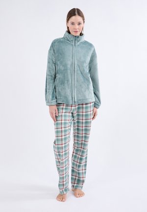 Mujer de pie descalza, vistiendo una chaqueta de forro polar azul claro y pantalones de pijama de cuadros en verde, rojo y blanco, sobre un fondo blanco.