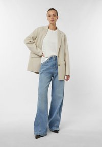 Beżowa marynarka narzucona na białą koszulkę, zestawiona z szerokimi niebieskimi dżinsami. Dżinsy mają jasne wybarwienie i lekko postrzępione krawędzie.