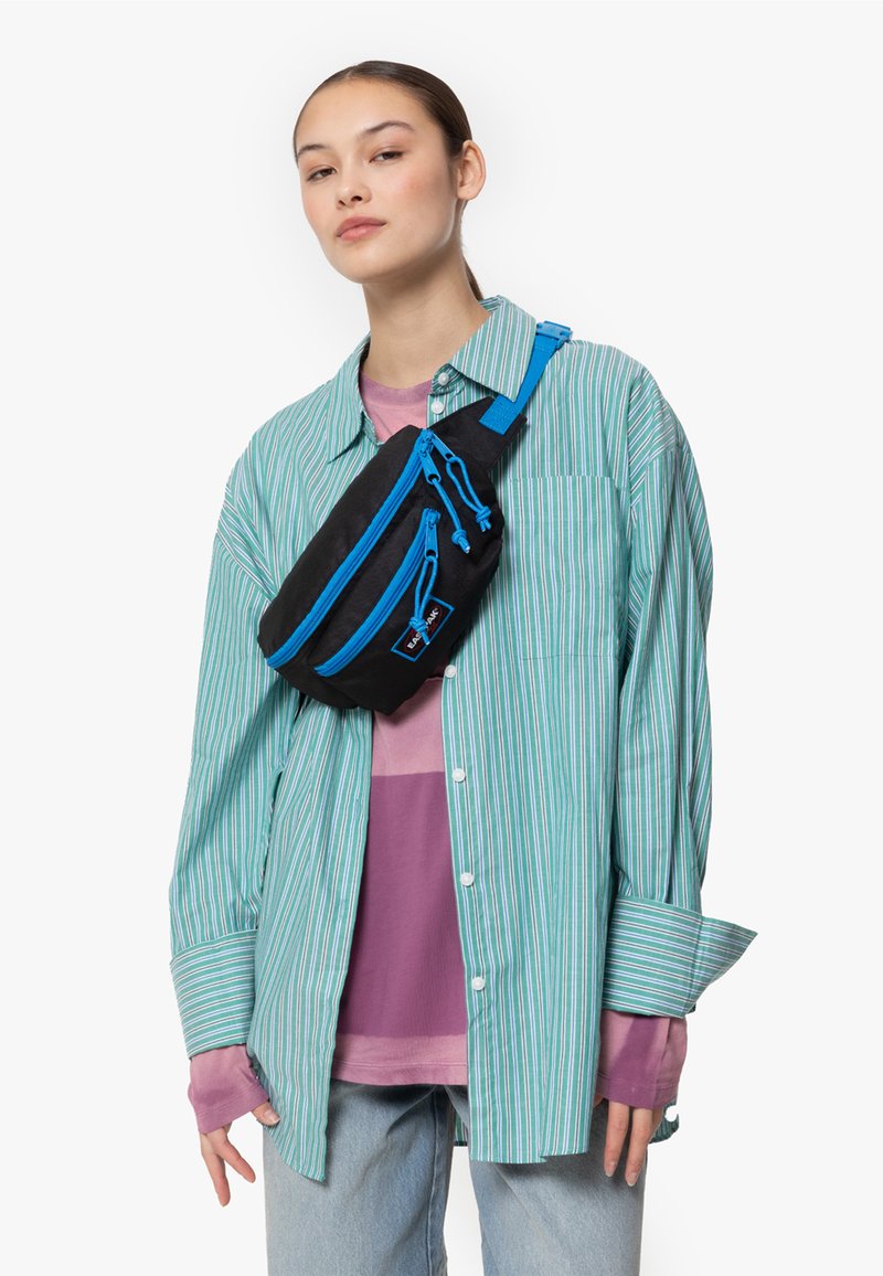 Černá crossbody taška s modrými zipy a akcenty, obdélníkový design, vyrobená z látky, nošena přes zelené pruhované tričko a vrstvené oblečení.