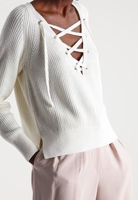 Witte gebreide trui met een diepe V-hals en een vetersluiting aan de voorkant. Voorzien van lange mouwen en een aansluitende geribde zoom.