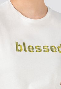 Vit bomull T-shirt med ordet "blessed" i texturerad grön och gul bokstäver. Rund halsringning, korta ärmar.
