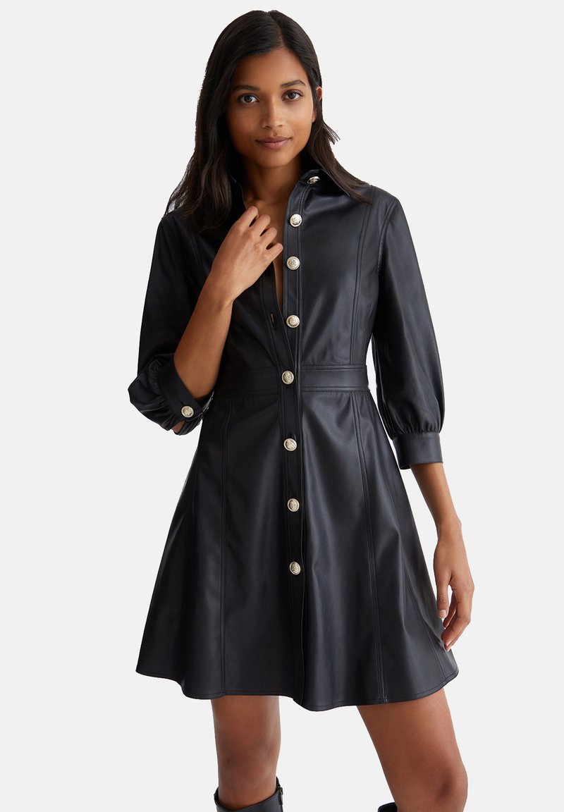 Schwarzes Kunstlederkleid mit Kragen, durchgehender Knopfleiste, Dreiviertelärmeln und ausgestelltem Rock. Mit großen silbernen Knöpfen.