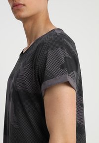 Černé tričko s maskáčovým vzorem, které má texturovaný design a šedé manžety. Tkanina vypadá lehce a neformálně.