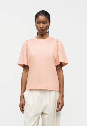 Γυναίκα με πλεξούδες, φορώντας ανοιχτό ροζ κοντομάνικη μπλούζα με ρυθμιζόμενους ώμους και άνετο λευκό παντελόνι, στέκεται μπροστά σε απλό φόντο.