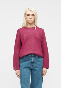 Ξανθιά γυναίκα που φοράει ένα ματζέντα πουλόβερ με μακριά μανίκια και μπλε τζιν, στέκεται μπροστά σε ένα απλό ανοιχτό γκρι φόντο, κοιτώντας μπροστά.