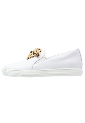 Sneaker slip-on bianco in pelle con un accento a forma di testa di leone dorata sul davanti, suola in gomma testurizzata bianca e design minimalista.
