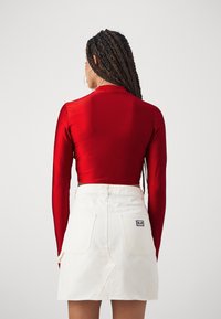 Κόκκινο κοντό τοπ με μακριά μανίκια σε γυαλιστερό ύφασμα, συνδυασμένο με λευκή τζιν φούστα που έχει μία μικρή μαύρη ετικέτα και δύο πίσω τσέπες.