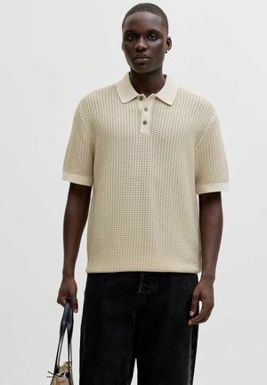 Junger Mann trägt ein beige strukturiertes kurzärmeliges Polohemd und schwarze Hose, hält eine schwarze Ledertasche in der linken Hand.