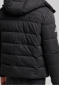 Czarna, puchowa kurtka z kapturem, z poziomymi quilted panelami. Wyhaftowane logo "SUPERDRY" na plecach i rękawie.