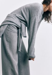 Person iført grå sweater og jeans, bøjende sig fremad mod ensartet lys baggrund, med sweaterærmer, der delvist dækker hænderne.