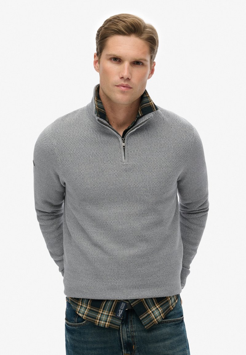 Lichtgrijze gebreide pullover met een halvemaan-rits kraag, gedragen over een groen en geruit overhemd, met lange mouwen en een gestructureerd patroon.