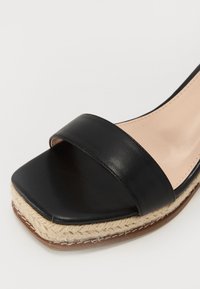 Sandalia deslizante de cuero negro con una tira ancha, forro interior beige y tacón en cuña envuelto en yute. Punta cuadrada y suela texturizada.