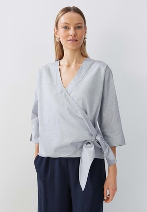 Top grigio chiaro avvolgente con scollatura a V, maniche a tre quarti, dettaglio con nodo in vita e una trama liscia, abbinato a pantaloni scuri.