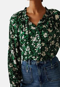 Kvinde iført en grøn blomsterbluse med lange ærmer og flæsekrave, stukket ned i højtaljede blå denimjeans.