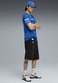Blå kortærmet skjorte med tekstureret design og logoer, sorte denimshorts, hvide sneakers og blå kasket. Har en synlig tatovering på benet.