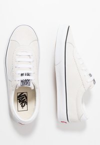 Baskets en daim blanc au design low-top, lacets blancs, semelles en caoutchouc texturé, accents noirs, et un logo Vans sur la languette et le talon.