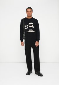 Fekete pulóver, amelyen két napszemüveget viselő figura grafikája látható, alatta a "KARL LAGERFELD" felirattal. Fekete cargo nadrággal és cipővel párosítva.