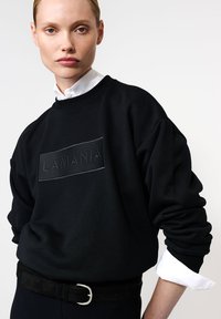 Juoda džemperis su gumbeliu ir rankovėmis, o priekyje įspaustas "LAMANIA" logotipas. Dėvimas ant balto marškinėlių su apykakle.