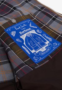 Plaid fabric in shades of grey, brown, and orange with a blue label featuring white graphics of a coat and accessories, including a tie and hat.