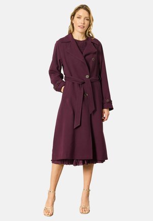Frau mit hellen Haaren, die einen dunkelvioletten Trenchcoat mit Gürtel und goldene Riemchenabsätze trägt, steht vor einem schlichten Hintergrund.