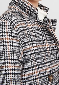 Kabát z vlněné směsi s kostkovaným vzorem v černé, bílé a oranžové barvě. Strukturovaný límec a detail zapínání na dva knoflíky.