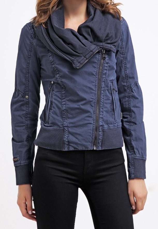 Femme portant une veste zippée bleu marine à col montant et un pantalon noir, debout avec les bras détendus le long du corps.