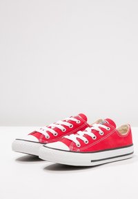 Une paire de baskets basses en toile rouge avec des semelles en caoutchouc blanc et des lacets blancs sur un fond blanc uni.