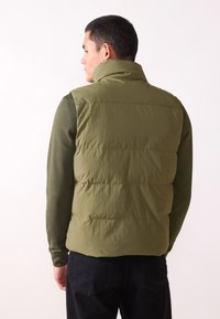 Olivenfarget puffer-vest med høy krave, sydd paneler og en glatt tekstur. Båret over en mørk langermet topp, sett fra baksiden.