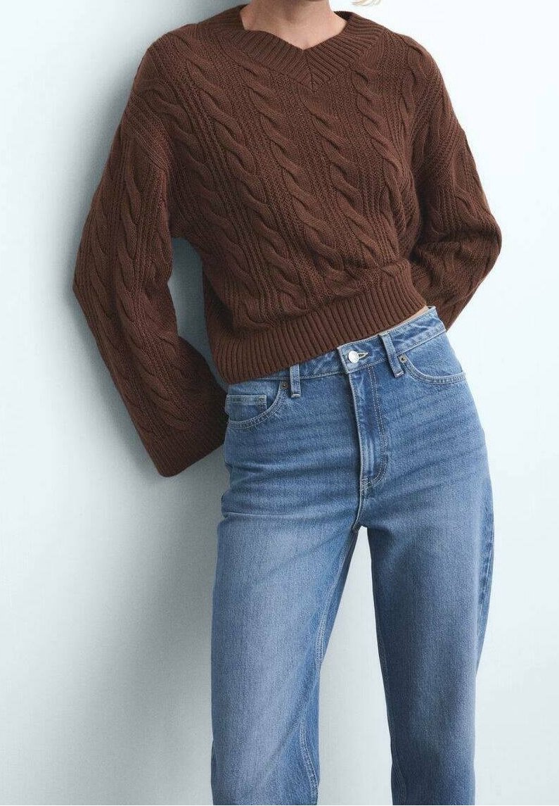 Personne portant un pull marron à torsades à manches longues et un jean bleu taille haute, debout contre un mur blanc.
