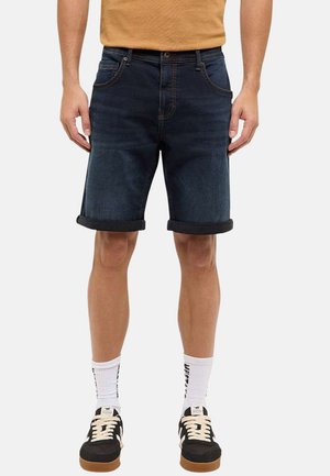 Mand iført mørkeblå denimshorts med opsmøg, hvide crew-strømper, sorte og beige sneakers samt en sennepsfarvet skjorte.
