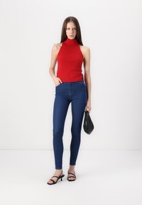 Rdeča brezrokavnik turtleneck majica, modre tesne kavbojke, črne sandale s peto in mala črna torbica; gladke teksture in prileganje.