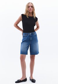 Top fitted nero senza maniche abbinato a pantaloni corti in denim blu con orlo sfrangiato. Scarpe nere flat completano l'outfit.