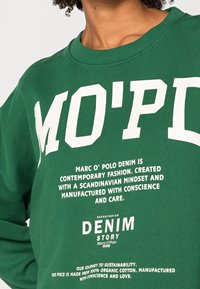 Vihreä puuvillainen collegepaita, jossa rohkeat valkoiset tekstit, pyöreä pääntie ja resorireunat hihoissa. Sisältää brändäys- ja kestävyystietoja.