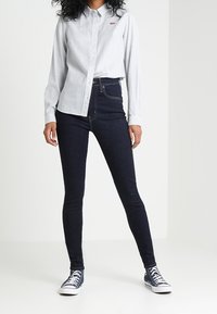 Camicia button-up a maniche lunghe di un blu chiaro con colletto; jeans skinny a vita alta in denim scuro; sneakers blu navy con suola e lacci bianchi.