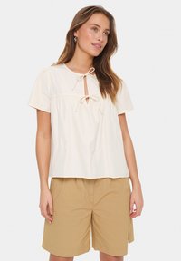 Camisetta corta beige chiaro con scollo rotondo, caratterizzata da un'apertura a chiave e dettaglio con fiocco, abbinata a pantaloni corti color khaki. Tessuto dalla consistenza morbida.
