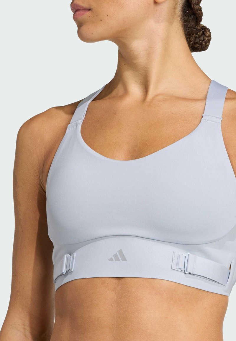Light grey sports bra with adjustable straps, a soft texture, and an elastic band featuring a logo at the center front under the bust.