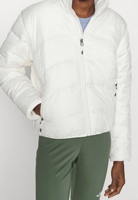 Osoba ubrana w białą kurtkę puchową North Face z zapięciem, z jedną ręką w kieszeni i zielone legginsy North Face.