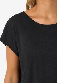 Tricou negru cu mâneci scurte și decolteu larg în formă de U, realizat dintr-un material moale și ușor, cu croială ușor lejeră și textură netedă.