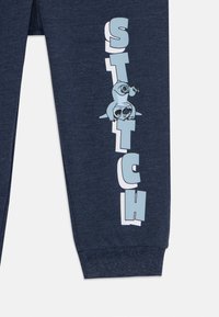 Tumši zili treniņbikses no mīksta auduma ar grafisku izdruku, kurā attēloti slothi, un lieliem baltajiem burtiem vārds "STITCH" sānos.